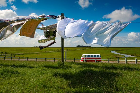 Rotweißer VW Bulli zwischen grünen Wiesen und einer Wäscheleine mit Wäsche im Vordergrund