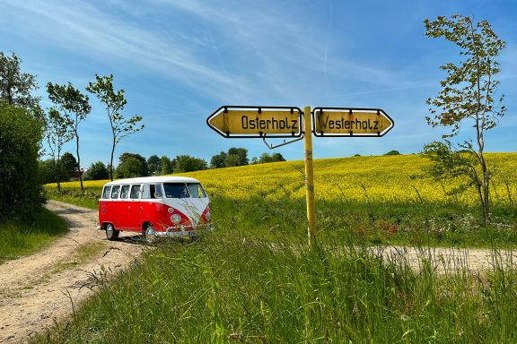 Rotweißer VW Bulli vor zwei Wegweisern und grünen Feldern